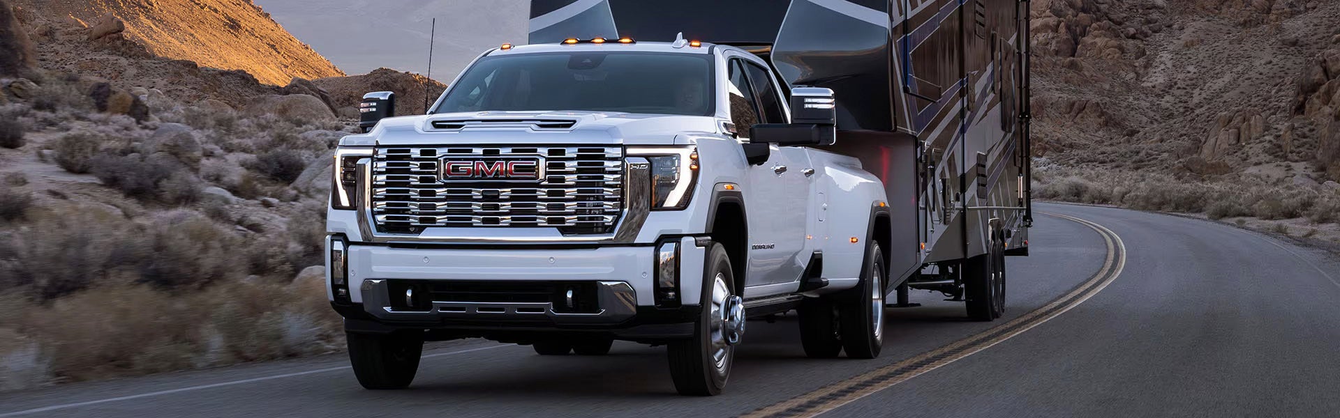 A white GMC Sierra HD Dually truck pulls a large fifth-wheel RV on a desert road with mountains in the background.