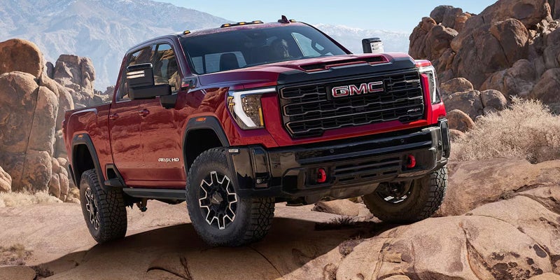 A red GMC Sierra pickup truck drives over rocky terrain, with mountains in the background.