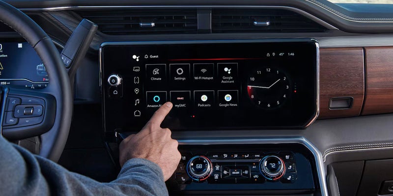 A hand touching the Amazon Alexa icon on a large, rectangular infotainment screen in a car. Applications, a clock, and climate controls are also visible.