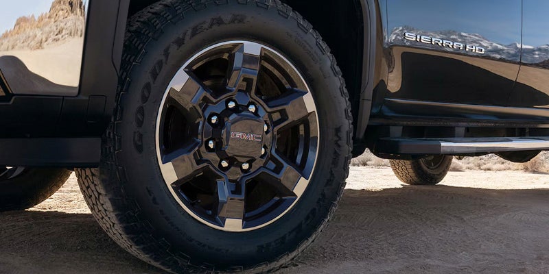 Close-up of a GMC Sierra HD truck's black and chrome wheel and all-terrain tire, with the truck's side visible.