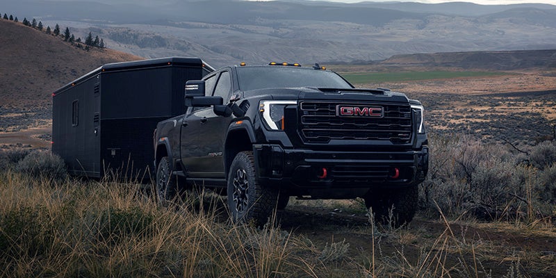 Black GMC Sierra 2500HD truck towing a black enclosed trailer on a dirt road through a dry, grassy landscape.